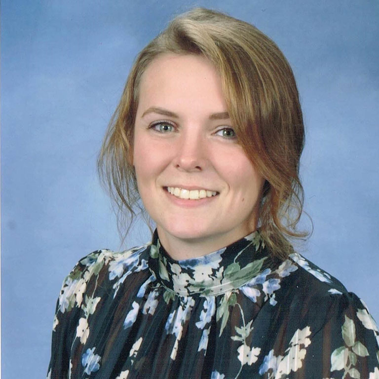 A headshot of Emma Bodiker, who wears a floral blouse and poses against a blue background.
