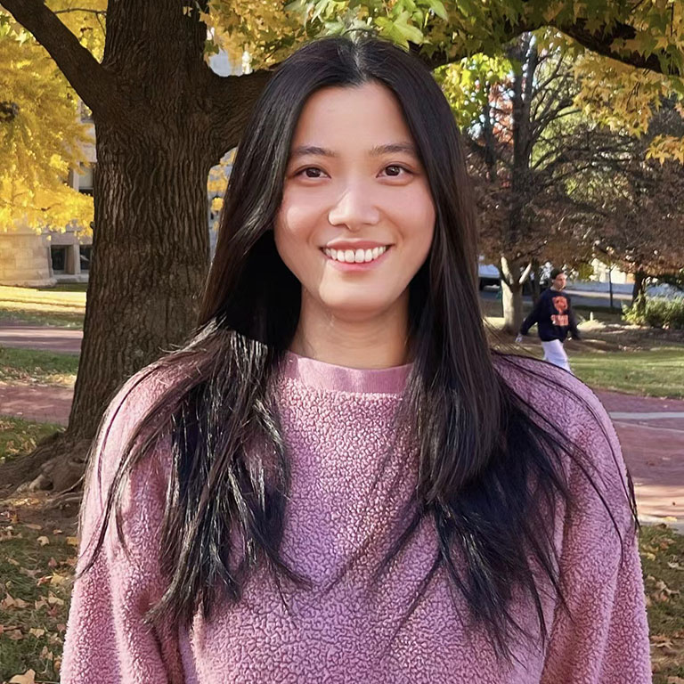A headshot of Qian Wu, who wears a pink sweater and poses outside.