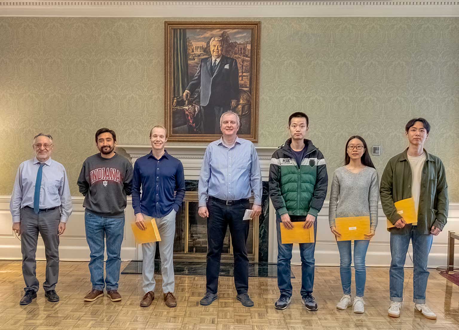 A group of graduate economics students pose in the Indiana Memorial Union, with a portrait of Herman B Wells above them.